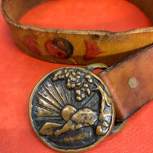 Op de afbeelding: Een vintage bruine leren riem met een messing gesp. De gesp heeft een zonsopgangontwerp met een boom en golven. De riem heeft een yin-yang symbool en rode accenten. De riem is ongeveer 3,8 cm breed.