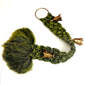 May include: A handmade, green crocheted item with a fluffy, moss-like end and a braided tail. The tail features decorative gold-colored metal accents and a circular ring at the end. The item is set against a white background.