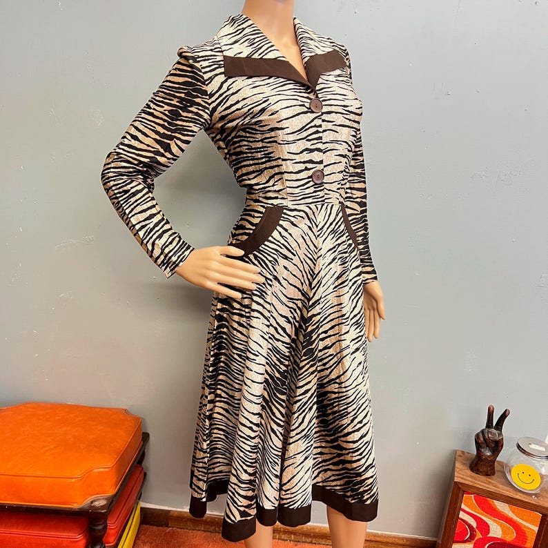 May include: A long-sleeved midi dress featuring a tan and black tiger stripe pattern. The dress has a pointed collar, a button-down front with two visible brown buttons, and solid brown trim along the collar, pocket flaps, and the hemline.