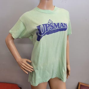 May include: A light green t-shirt with the word "UPSMAN" printed in dark blue across the chest. The shirt has short sleeves and a crew neck. The text is slightly distressed, giving it a vintage look.