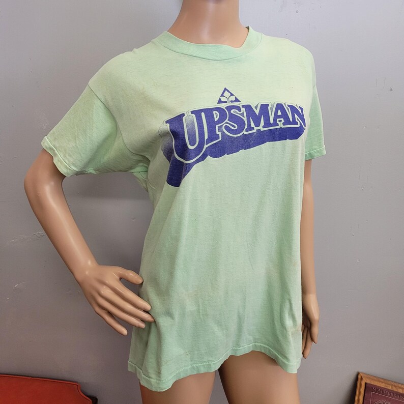 May include: A light green t-shirt with the word "UPSMAN" printed in dark blue across the chest. The shirt has short sleeves and a crew neck. The text is slightly distressed, giving it a vintage look.