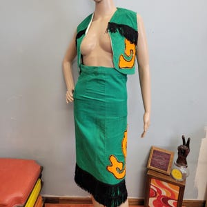 May include: A vintage green two-piece outfit consisting of a vest and a skirt. The vest has black fringe and orange applique. The skirt has orange applique and black fringe at the hem. The outfit is displayed on a mannequin.