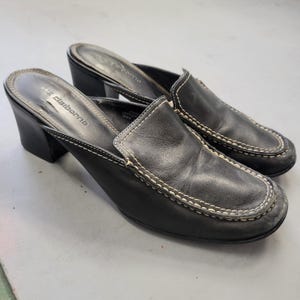 May include: A pair of black leather mules with a block heel and a rounded toe. The shoes feature prominent white contrast stitching along the vamp and the perimeter of the footbed. The Liz Claiborne brand name is embossed on the black insole.