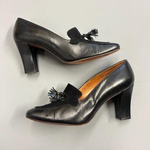 May include: A pair of black leather heeled loafers with a square toe and a suede vamp. Each shoe features a tassel detail and a stacked heel. The shoes are displayed on a light gray surface.