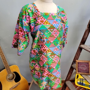 Könnte beinhalten: Ein farbenfrohes Vintage-Kleid mit quadratischem Ausschnitt und Dreiviertelärmeln. Das Kleid hat ein lebendiges geometrisches Muster mit rosa, grünen, blauen und braunen Formen, darunter Herzen und Blumen. Das Kleid wird auf einer Schaufensterpuppe präsentiert.