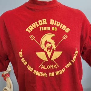 May include: Red t-shirt with gold lettering and graphics. The text reads "TAYLOR DIVING TEAM 89" above a helmet and trident design. Below the design, the text reads "ALOHA" and "NO SEA TOO ROUGH; NO MUFF TOO TOUGH."