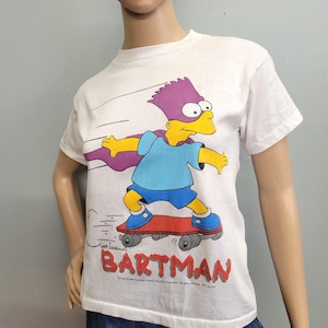 May include: White t-shirt featuring Bart Simpson as Bartman, skateboarding. Bart is depicted in a blue shirt, purple mask and cape, and blue shoes. The word "BARTMAN" is printed in red at the bottom. The shirt is a vintage item.