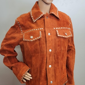 May include: A burnt orange suede jacket adorned with silver studs. The jacket has a traditional collar, button fastening, and two chest pockets. Silver studs are placed along the collar, pockets, cuffs, and shoulders, providing a decorative touch to the jacket.