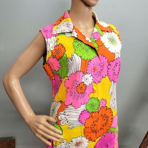 Puede incluir: Un vestido sin mangas con cuello que presenta un vibrante estampado floral multicolor en tonos amarillo brillante, rosa intenso, verde lima y naranja. La prenda tiene un cierre frontal de botones pequeños de color naranja y tela plisada.