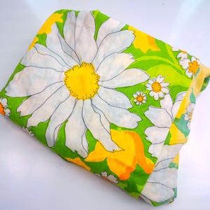 May include: Folded fabric with a large daisy floral pattern. The fabric features white daisies with yellow centers against a green background. Smaller daisies and yellow accents are also present. The fabric is folded, showing the pattern on the top.