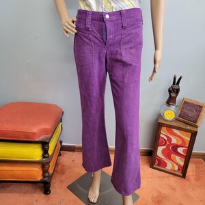 May include: Purple flared pants with front pockets and a button closure. The pants are displayed on a mannequin. The pants have a high waist and a flared leg. The pants are made of a textured fabric.