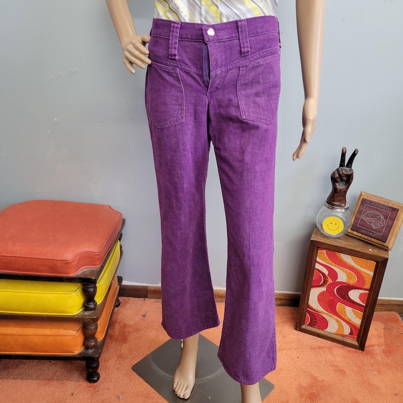 May include: Purple flared pants with front pockets and a button closure. The pants are displayed on a mannequin. The pants have a high waist and a flared leg. The pants are made of a textured fabric.