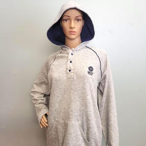 May include: A light gray hooded sweatshirt with a navy blue interior lining. The sweatshirt has a front pocket, a button-up neckline, and a small logo on the chest. Black trim outlines the hood and sleeves.