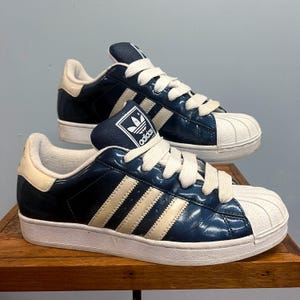 Peut inclure: Une paire de baskets Adidas Superstar bleu marine et crème. Les baskets ont des lacets blancs, un bout de pied blanc et le logo Adidas sur la languette. Les trois bandes emblématiques sont de couleur crème.