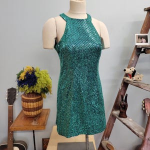 May include: A teal sequined halter dress displayed on a mannequin. The dress is sleeveless and has a high neckline. The sequins create a shimmering effect. The dress is a mini length.