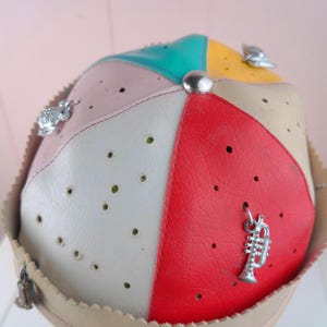 Op de afbeelding: Een veelkleurige patchwork-stijl hoed met rode, witte, gele en turquoise panelen. De hoed is versierd met zilveren bedels en een zilveren knoop. De rand is lichtbeige.