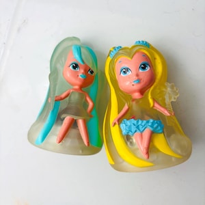 Könnte beinhalten: Zwei kleine Kunststofffiguren mit stilisierten Charakteren, großen Augen und buntem Haar. Eine Figur hat durchscheinendes mintgrünes Haar und ein graues Kleid, die andere gelbes Haar, einen blauen Rüschenrock und einen sternförmigen Zauberstab.