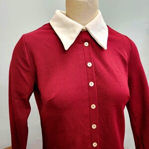 May include: A vintage burgundy dress with a cream-colored collar. The dress features long sleeves and a button-down front with six white buttons. The fabric has a ribbed texture, and the dress is displayed on a mannequin.