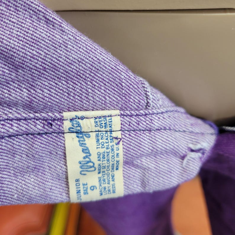May include: Close-up of purple denim fabric with a Wrangler tag. The tag reads "JUNIOR SIZE 9" and includes washing instructions. The denim is a solid, vibrant purple color.