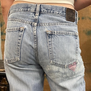 Puede incluir: Vaqueros de mezclilla azul claro con un aspecto descolorido y desgastado. Los vaqueros tienen un parche en el bolsillo trasero con un diseño de corazón rosa. Los vaqueros tienen un parche de cuero en la cintura trasera con el texto "WORLDWIDE" en un círculo.