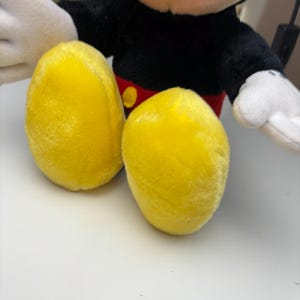 May include: Close-up of a plush toy featuring bright yellow feet. The toy has a black body, red shorts, and white gloves. The feet are oval-shaped and made of a soft, textured material.