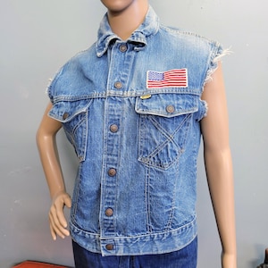 May include: A light blue denim waistcoat featuring a frayed sleeveless design, a classic collar, and a front button closure. The garment includes two chest pockets with button flaps and a rectangular embroidered patch of the American flag on the chest.