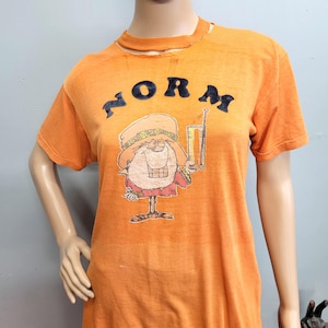 May include: An orange short-sleeved t-shirt featuring a distressed graphic of a cartoon character wearing a hat and holding a tall glass. The word NORM is printed in dark, textured letters above the character. The neckline has a frayed, worn edge.
