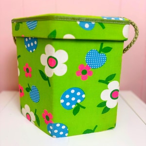 Könnte beinhalten: Eine grüne Box mit Deckel und geflochtenem Griff, verziert mit einem Blumenmuster. Das Design umfasst weiße Blumen mit rosa Zentren, blaue gepunktete Fruchtformen und rosa Blumen auf einem hellgrünen Hintergrund. Der Deckel ist grün eingefasst.