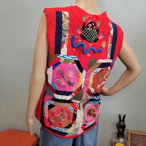 May include: A red quilted vest with a patchwork design. The vest features embroidered details, including a black creature with red eyes, a blue wavy line, and various colourful geometric shapes. The vest is sleeveless and has a round neckline.