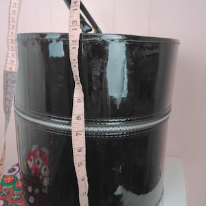 May include: A black, glossy, cylindrical handbag with a silver zipper and a black handle. The bag is approximately 12 inches tall. A measuring tape is next to the bag, showing the height.