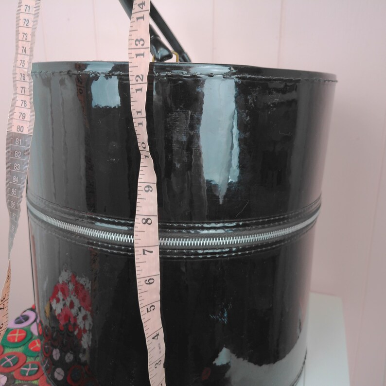 May include: A black, glossy, cylindrical handbag with a silver zipper and a black handle. The bag is approximately 12 inches tall. A measuring tape is next to the bag, showing the height.