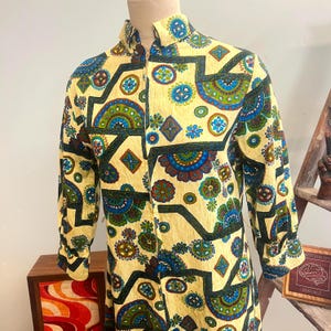 Op de afbeelding: Een vintage jurk met hoge kraag en lange mouwen, gepresententeerd op een mannequin. De crèmekleurige jurk heeft een herhalend patroon van blauwe, groene en bruine bloemen en geometrische vormen. De jurk heeft een ritssluiting.
