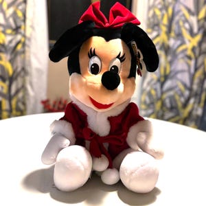 Pode incluir: Uma boneca de pel&uacute;cia da Minnie Mouse, com orelhas pretas, um la&ccedil;o vermelho e uma roupa vermelha e branca. A boneca est&aacute; sentada com as pernas estendidas e os bra&ccedil;os ao lado. A boneca tem um nariz preto, olhos grandes e uma boca vermelha sorrindo.