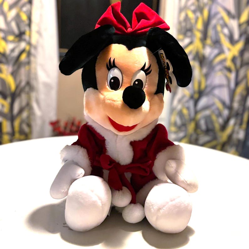Pode incluir: Uma boneca de pel&uacute;cia da Minnie Mouse, com orelhas pretas, um la&ccedil;o vermelho e uma roupa vermelha e branca. A boneca est&aacute; sentada com as pernas estendidas e os bra&ccedil;os ao lado. A boneca tem um nariz preto, olhos grandes e uma boca vermelha sorrindo.