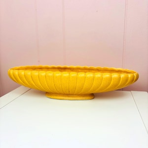 May include: A large, oval-shaped, yellow ceramic planter with a textured surface and a scalloped rim. The planter sits on a small, round base. The background is a light pink wall.
