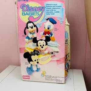 May include: Vintage Disney Babies toy box featuring plush figures of Baby Mickey Mouse, Baby Minnie Mouse, Baby Goofy, and Baby Donald Duck. The box is pink with the Disney Babies logo and text. The box is from 1984.