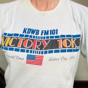 Puede incluir: Una camiseta blanca de manga larga con un gráfico que muestra el texto KDWB FM 101 VICTORY 10K. El diseño incluye un borde de tira de película azul, una bandera estadounidense roja, blanca y azul, y el texto Memorial Drive Labor Day, 1984.