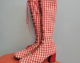 Puede incluir: Un par de botas hasta la rodilla con un estampado de cuadros rojos y blancos. Las botas tienen cordones rojos, tacones de bloque rojos y ojales de bronce. Las botas se exhiben sobre una superficie naranja.