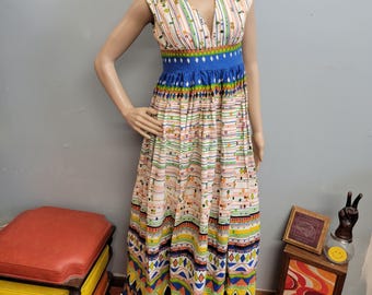 May include: A sleeveless, floor-length dress with a V-neckline. The dress features a cream-colored background with vertical stripes in various colors. A blue band accents the waist, and the skirt has a patterned design with geometric shapes and vibrant colors.