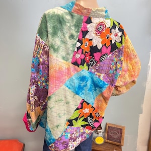 May include: A colourful patchwork jacket with a quilted design. The jacket features a mix of floral, tie-dye, and geometric patterns in pink, blue, green, and orange. The sleeves are long, and the collar is a contrasting colour. The jacket is made of various fabric patches.