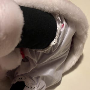 Pode incluir: Close-up de uma fantasia de Papai Noel de pel&uacute;cia branca com uma borda branca fofa. A fantasia inclui uma perna preta e uma saia branca com detalhes de renda. Um detalhe vermelho &eacute; vis&iacute;vel.