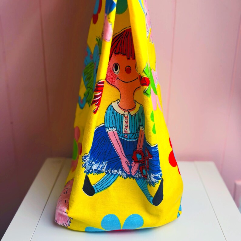 May include: A yellow tote bag with a vintage doll illustration. The doll has red hair, a blue dress, and black shoes. The bag's background is decorated with colourful floral and abstract shapes.