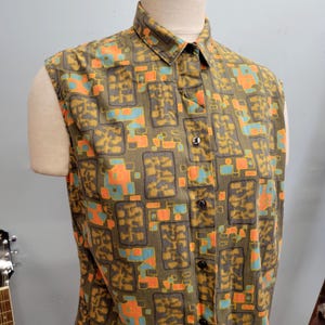Peut inclure: Chemise sans manches à motif géométrique vert olive, orange et bleu. La chemise a un col et des boutons foncés. Le tissu semble être du coton ou du lin léger. La chemise est boutonnée.