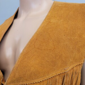 May include: Mustard yellow suede vest with a V-neck and fringed hem. The vest is displayed on a mannequin. The suede has a soft, textured appearance. The fringing is along the bottom edge of the vest.