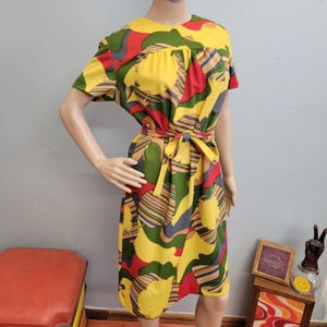 Könnte beinhalten: Ein Vintage-Kleid in knielanger Länge mit einem farbenfrohen, abstrakten Muster. Das Kleid hat kurze Ärmel, einen Bindegürtel und eine lebendige Mischung aus gelben, roten, grünen und grauen Formen. Das Kleid wird auf einer Schaufensterpuppe präsentiert.