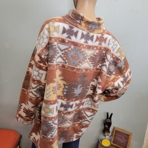 K&ouml;nnte beinhalten: Ein Fleece-Pullover mit hohem Kragen. Der Pullover hat ein vom S&uuml;dwesten inspiriertes Muster in Braun-, Creme-, Gelb- und Blaut&ouml;nen. Das Design umfasst geometrische Formen und stilisierte Motive.