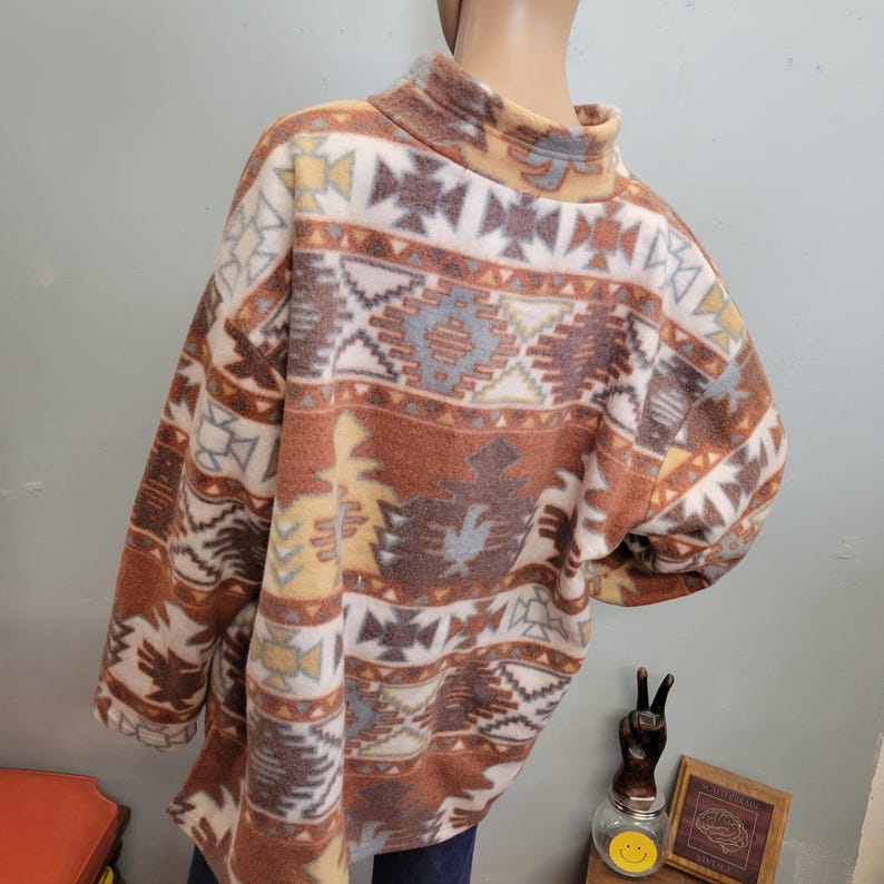 K&ouml;nnte beinhalten: Ein Fleece-Pullover mit hohem Kragen. Der Pullover hat ein vom S&uuml;dwesten inspiriertes Muster in Braun-, Creme-, Gelb- und Blaut&ouml;nen. Das Design umfasst geometrische Formen und stilisierte Motive.