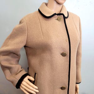 May include: A tan wool coat with black trim and gold buttons. The coat has a classic design with a rounded collar and a tailored fit. The black trim outlines the collar, cuffs, and front closure. The gold buttons add a touch of elegance.