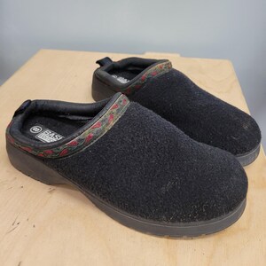May include: A pair of black Easy Spirit clogs. The clogs have a textured, fuzzy upper and a dark sole. The collar is trimmed with a patterned band of red, green, and brown. The interior is lined with a soft material.