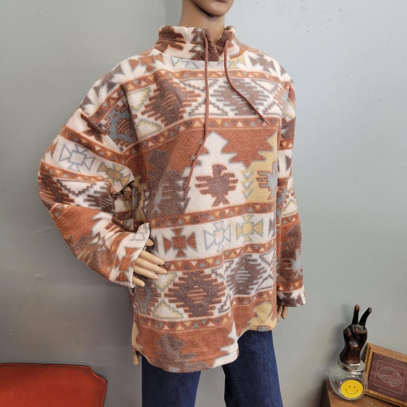 K&ouml;nnte beinhalten: Ein Fleece-Pullover mit hohem Kragen und Kordelzug. Der Pullover hat ein Muster im S&uuml;dwest-Stil in Braun-, Creme- und Graut&ouml;nen. Das Design umfasst geometrische Formen und stilisierte Vogelmotive. Die &Auml;rmel sind lang, und das Kleidungsst&uuml;ck wird auf einer Schaufensterpuppe pr&auml;sentiert.